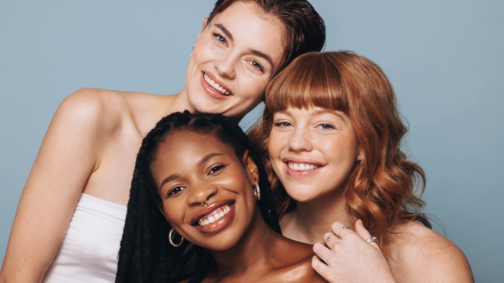 Tri žene različitih tonova kože poziraju nasmejane ispred plave pozadine - slika oslikava značaj melanina u nezi kože